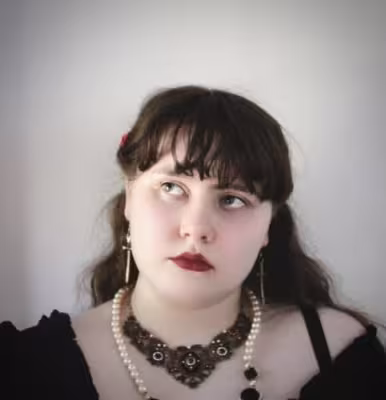 Riley stands in front of a grey background wearing gothic jewellery and a black dress.